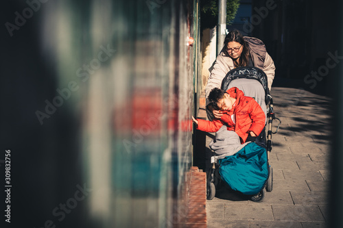 una mujer (la madre) lleva a su hijo con pluridiscapacidad de paseo por la calle en invierno con cochecito adaptado (silla de ruedas), el niño se acerca al muro para tocarlo con la mano, estimulo sens