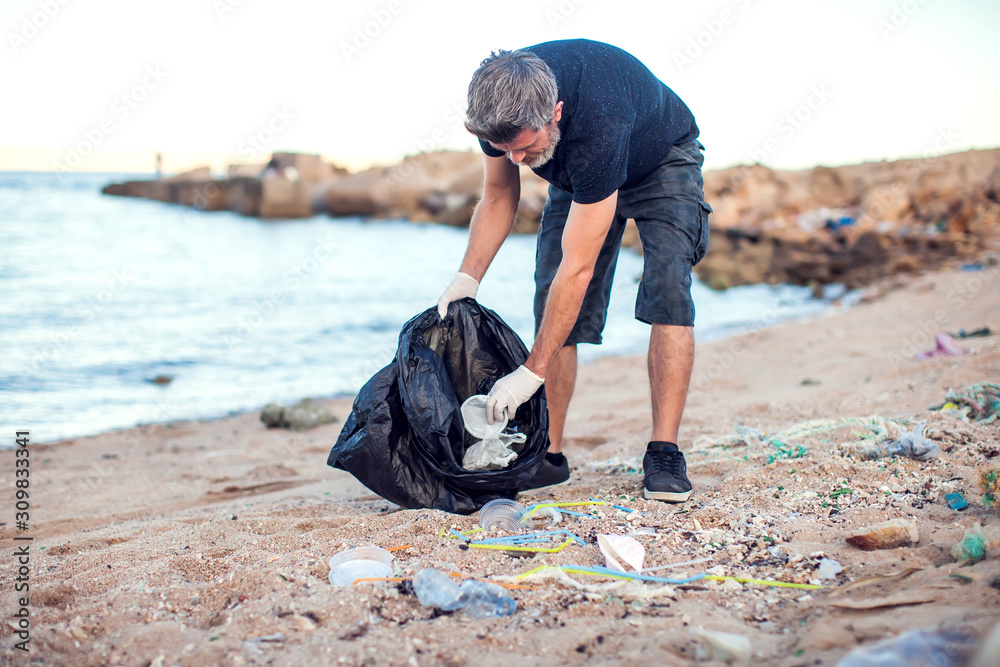 Man with white gloves and big black package collecting garbage on the beach. Environmental protection and planet pollution concept Man with white gloves and big black package collecting garbage on the beach. Environmental protection and planet pollution concept