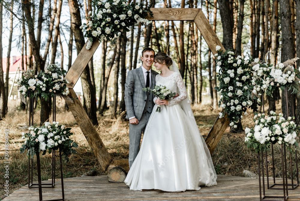 Attractive couple celebrating their wedding in forest. Portrait of young happy groom and bride in wedding clothes standing together, holding hands and looking at each other near arch