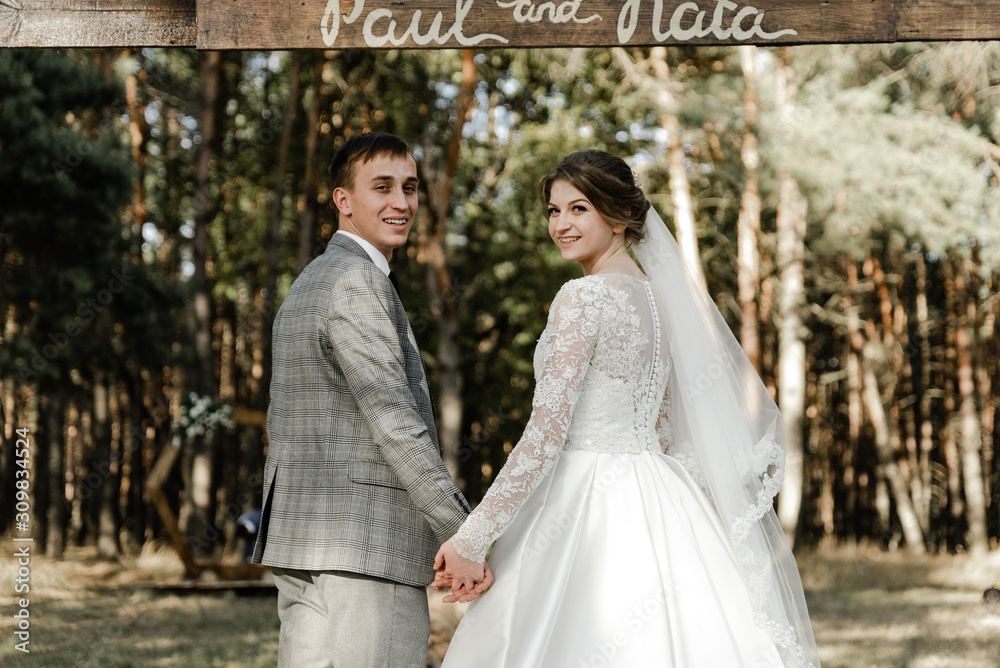 Attractive couple celebrating their wedding in forest. Portrait of young happy groom and bride in wedding clothes standing together, holding hands and looking at each other near wedding photozone