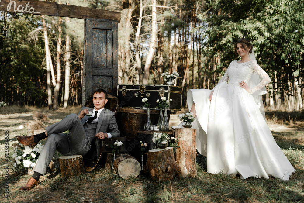 Attractive couple celebrating their wedding in forest. Portrait of young happy groom and bride in wedding clothes standing together, holding hands and looking at each other near wedding photozone