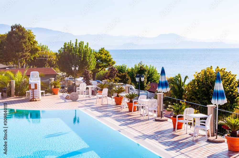Outdoor pool with vibrant crystal water, parasols and deck chairs located on the coast of Garda with lake, hills and sun on background