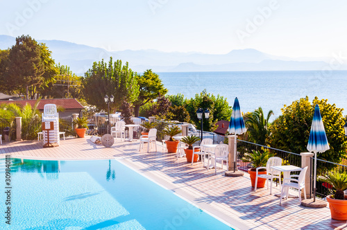 Outdoor pool with vibrant crystal water, parasols and deck chairs located on the coast of Garda with lake, hills and sun on background