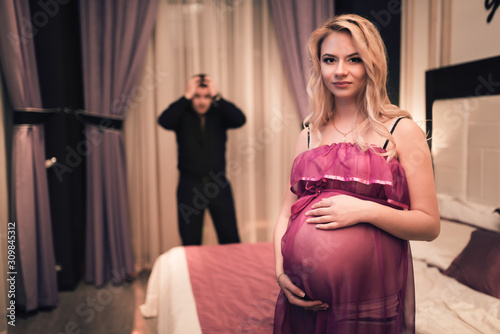 Young pregnant beautiful woman holds her stomach while standing against a blurred tired husband clutching his head. Concept of vagaries of a pregnant woman photo
