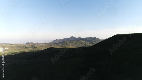 Aerial view of the small settlement in green hills with high mountain chain and blue sea on the background against blue sky. Shot. Beautiful colorful landscape.