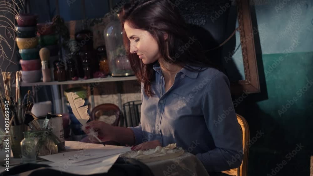 Focused woman is writing a letter with bird's feather and ink while sitting at her nice workshop.