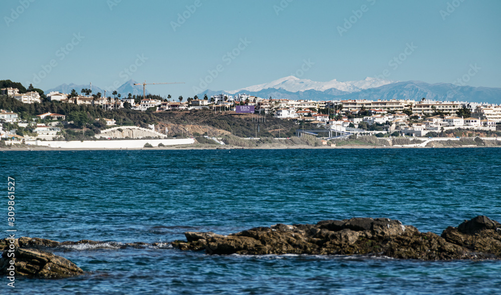 Malaga coastline seen from La Cala de Mijas beach on a sunny winter afternoon with the white peaks of Sierra Nevada mountain range at the horizon.