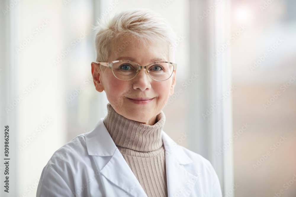 Head and shoulders portrait of friendly female doctor smiling at camera while standing against window in clinic, copy space Head and shoulders portrait of friendly female doctor smiling at camera while standing against window in clinic, copy space