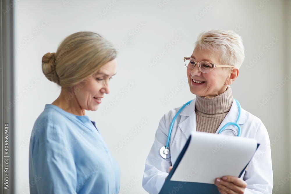 Waist up portrait of mature female doctor smiling cheerfully while talking to senior woman preparing her for medical procedures in clinic, copy space Waist up portrait of mature female doctor smiling cheerfully while talking to senior woman preparing her for medical procedures in clinic, copy space