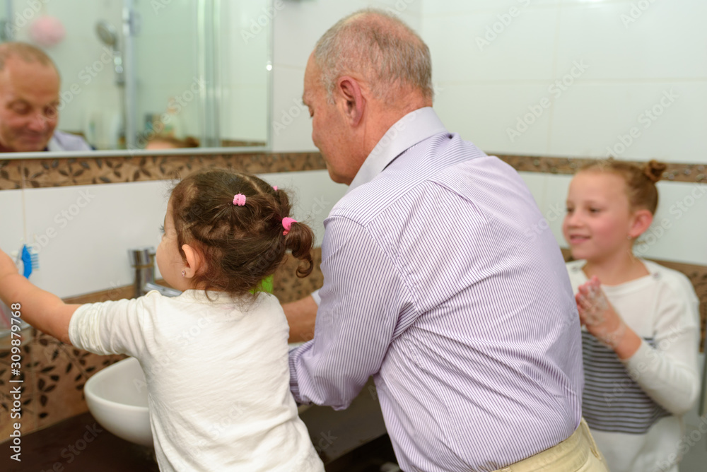Little happy toddler girls and grandfather in bathroom washing hands. Healthy, Child's and People's Hygiene concept. Selective focus on little brunette girl. Little happy toddler girls and grandfather in bathroom washing hands. Healthy, Child's and People's Hygiene concept. Selective focus on little brunette girl.