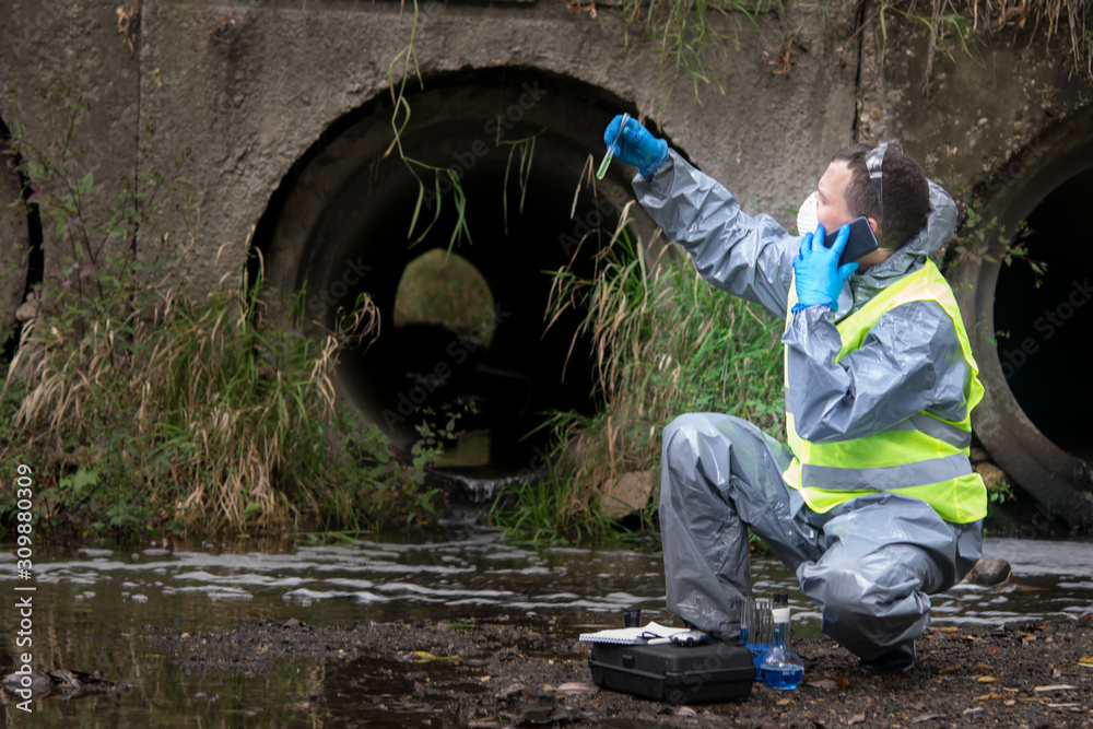 scientist in protective suit, gloves and mask, holding a vial of liquid and is calling on mobile phone user to report results of samples from the river, amid the purifying pipe scientist in protective suit, gloves and mask, holding a vial of liquid and is calling on mobile phone user to report results of samples from the river, amid the purifying pipe