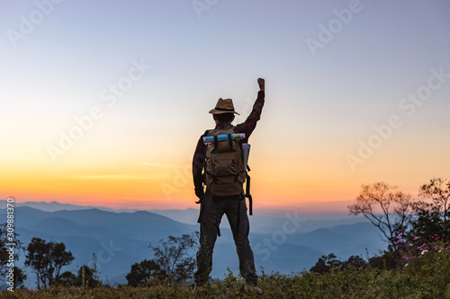 Young man with backpack standing with raised hands on top of mountain at sunset