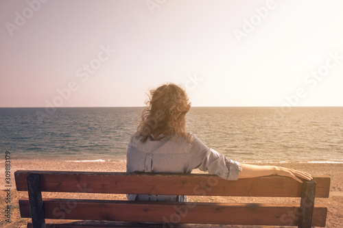 Rear view of young woman wearing blue casual dress sitting on a bench while looking away to horizon and sunset over sea