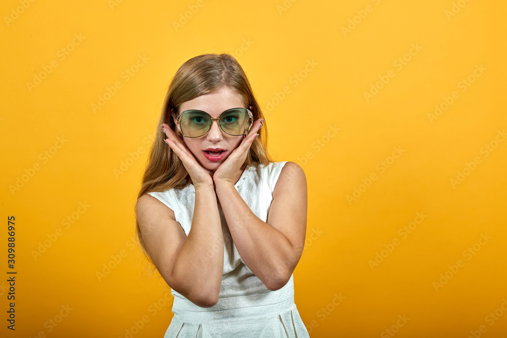 Charming blonde young woman having casual sunglasses, holding hand over chin, looking shocked over isolated orange background in white shirt. Lifestyle concept Charming blonde young woman having casual sunglasses, holding hand over chin, looking shocked over isolated orange background in white shirt. Lifestyle concept