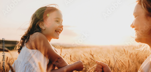 Side view portrait of a happy little girl laughing while looking at her mother in a field of wheat against sunset. photo