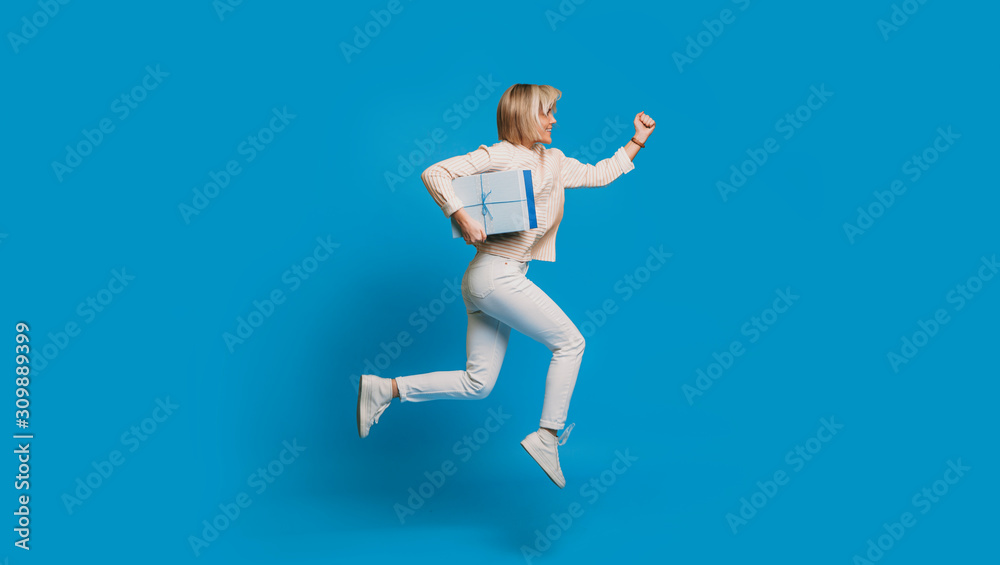 Side view portrait of a beautiful young woman running with a blue gift box against a blue background. Fast delivery concept.
