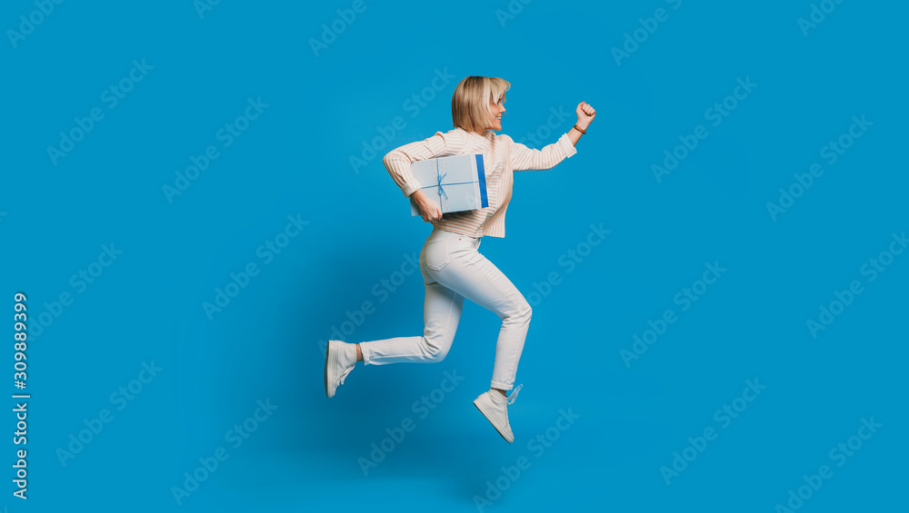 Fototapeta premium Side view portrait of a beautiful young woman running with a blue gift box against a blue background. Fast delivery concept.
