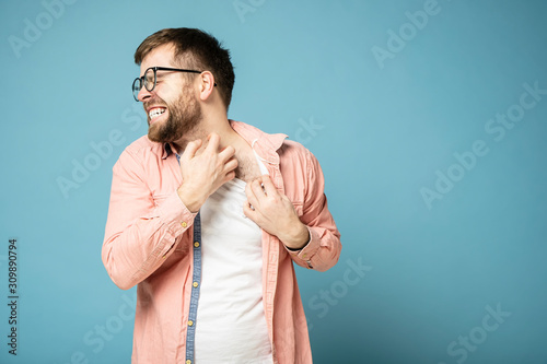 Tense bearded man in glasses scratches itchy skin on neck with his hand, painfully grins teeth and closes eyes. Blue background. photo
