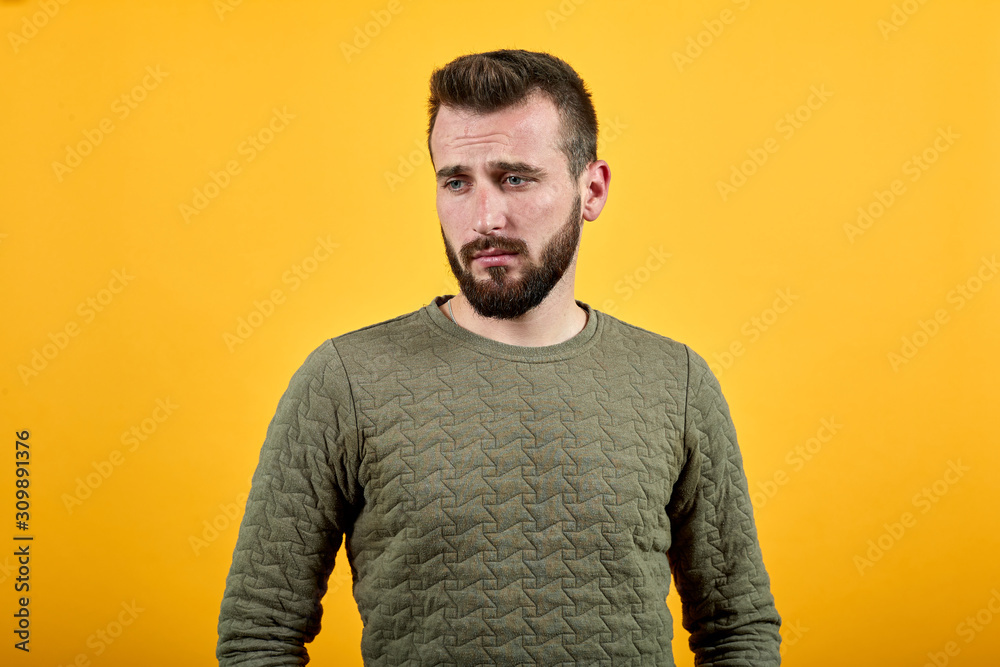 Serious handsome caucasian young man over isolated orange background looking down, keeping hand on waist wearing fashion green sweater Serious handsome caucasian young man over isolated orange background looking down, keeping hand on waist wearing fashion green sweater
