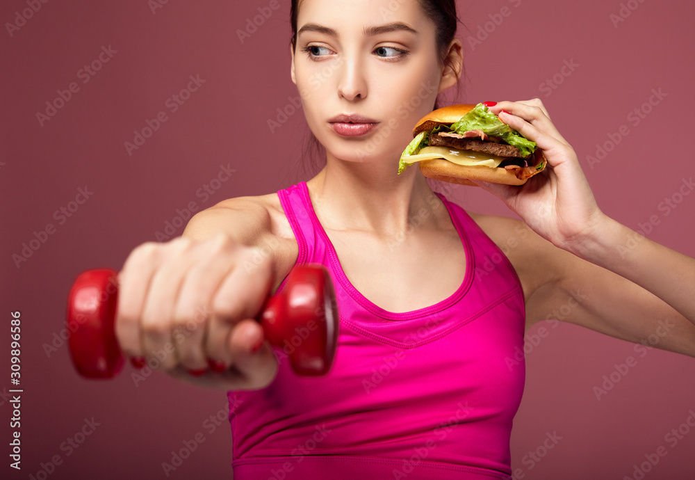 Athletic girl with a beautiful body holds a burger and dumbbells. Athletic girl with a beautiful body holds a burger and dumbbells.