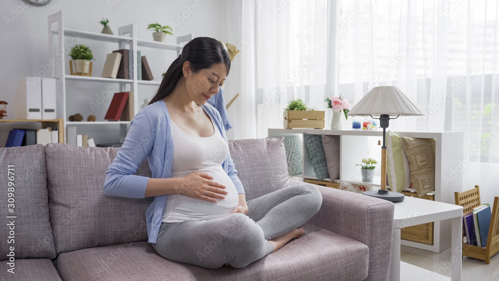Smiling asian chinese pregnant woman sitting in comfort sofa looking at big stomach with hands holding. young elegant future motherhood crossed legs relax in couch in living room at bright apartment Smiling asian chinese pregnant woman sitting in comfort sofa looking at big stomach with hands holding. young elegant future motherhood crossed legs relax in couch in living room at bright apartment