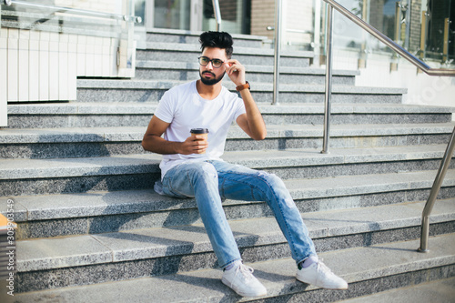 Young indian man in glasses drink coffee sitting on the stairs in the street