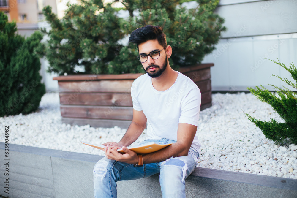 Obraz premium Young indian man in glasses read notebook sitting on the bench in the street