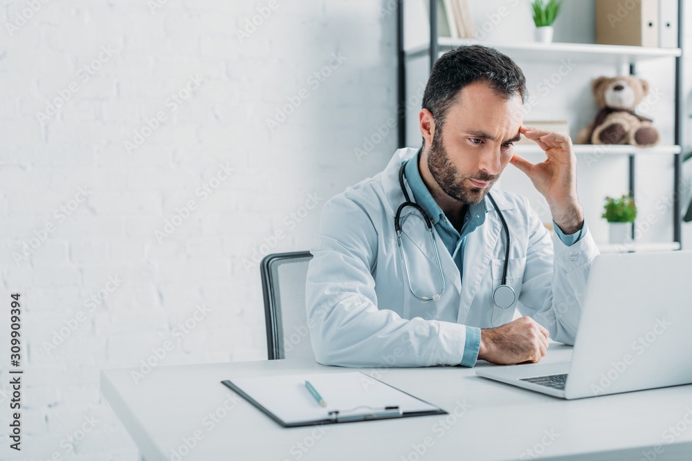 thoughtful doctor looking at laptop while sitting at workplace