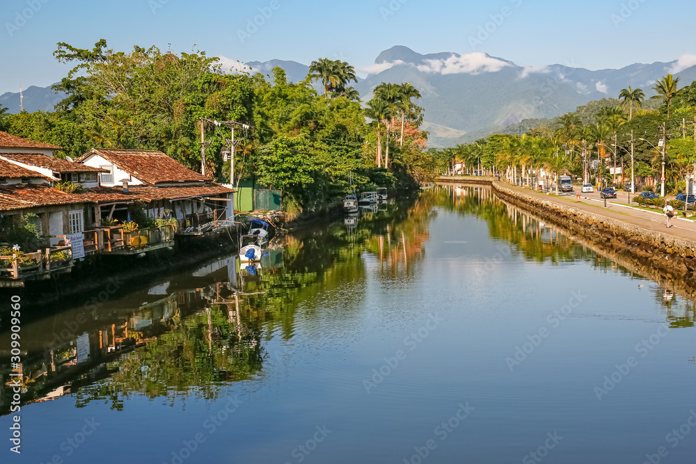 View of river Perequê-Açu with houses, vegetation and rainforest mountains in background on a sunny day, reflections on the water in Unesco World Heritage town Paraty, Brazil