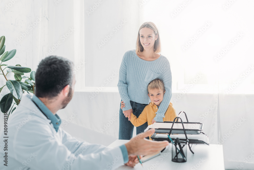 smiling mother hugging cheerful son while looking at pediatrist in hospital