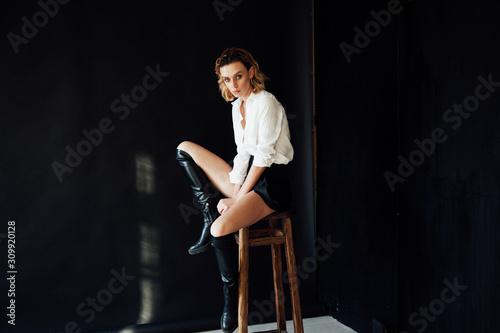 Beautiful fashionable blonde woman in a white blouse and black shorts with boots on a black background