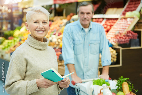 Waist up portrait of modern senior woman holding shopping list and smiling at camera while enjoying grocery shopping in supermarket, copy space