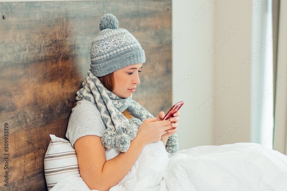 Fototapeta premium ill young female in hat and scarf lying on bed suffering from illness, disease, watching video on smartphone, have rest, have sick leave at home