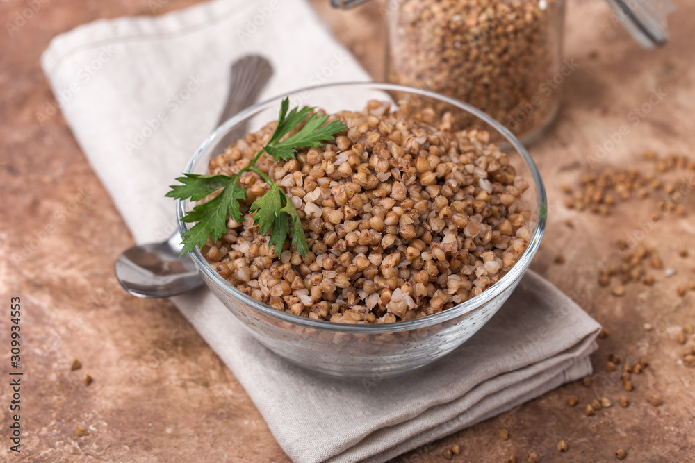 Cooked buckwheat in a glass, transparent bowl, parsley leaf, linen napkin, jars for storing cereals, brown background Cooked buckwheat in a glass, transparent bowl, parsley leaf, linen napkin, jars for storing cereals, brown background