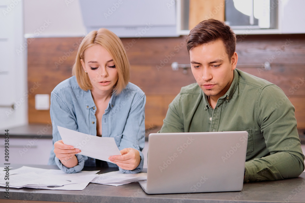 young caucasian couple search bills and expenses on mortgage using laptop at home, sit together and look at screen of laptop, with documents on table young caucasian couple search bills and expenses on mortgage using laptop at home, sit together and look at screen of laptop, with documents on table