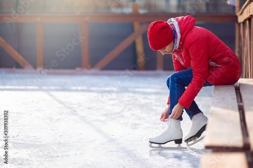 Girl in a red jacket sits on a bench on an open ice rink and laces figure skates