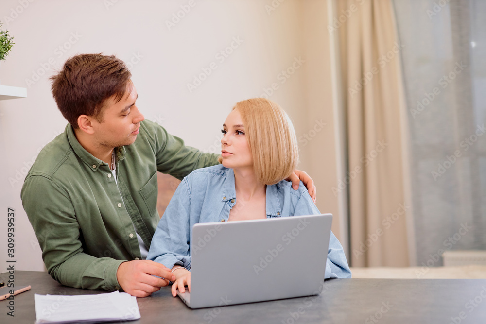 young caucasian woman busy with financial problems at home using laptop , her husband attentively look at her and listen. young family solve problems young caucasian woman busy with financial problems at home using laptop , her husband attentively look at her and listen. young family solve problems