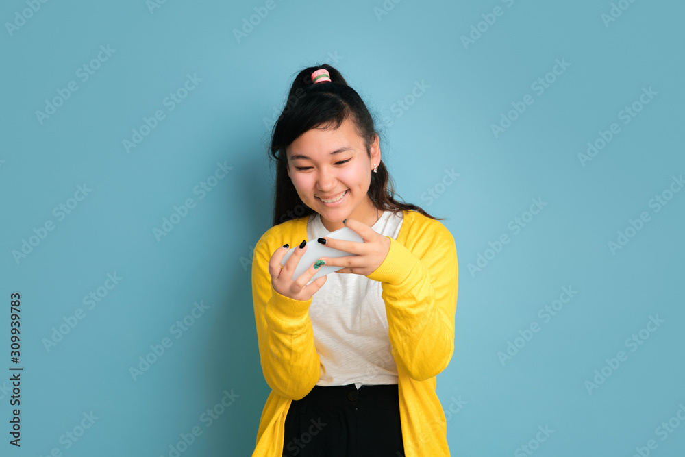 Asian teenager's portrait isolated on blue studio background. Beautiful female brunette model with long hair. Concept of human emotions, facial expression, sales, ad. Using phone, smiling, happy win. Asian teenager's portrait isolated on blue studio background. Beautiful female brunette model with long hair. Concept of human emotions, facial expression, sales, ad. Using phone, smiling, happy win.