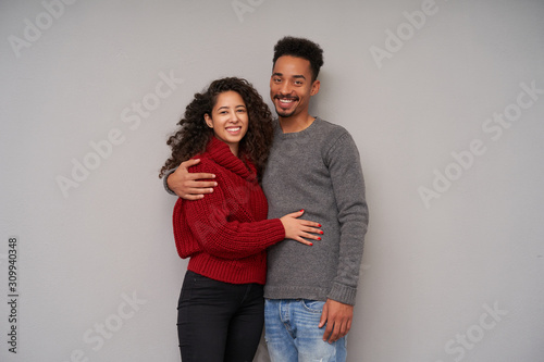 Young attractive dark skinned brunette couple hugging gently each other and looking happily at camera with wide sincere smiles, wearing knitted woolen sweaters and jeans over grey background