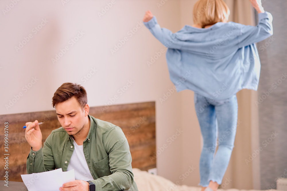 Fototapeta premium tired busy man sit on table working on laptop, checking documents while his wife jumping on bed, in casual wear