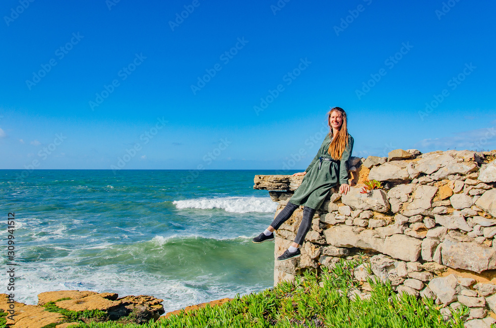 Woman dreamily looks at the Atlantic ocean and smiles, sits on the edge of a rocky shore, the wind blows hair, makes waves. Copy space.