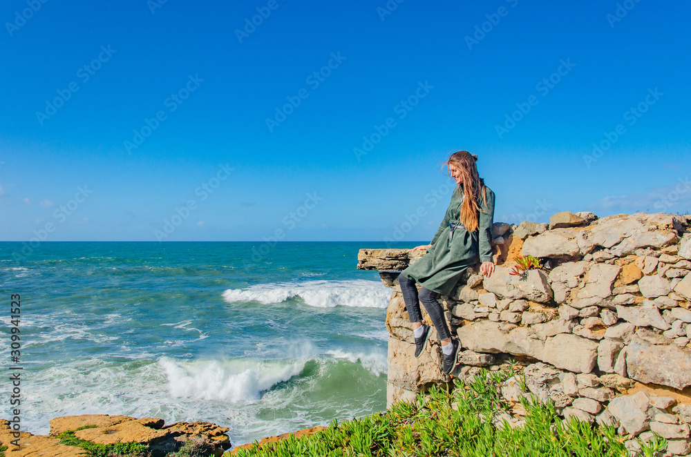 Obraz premium Woman dreamily looks at the Atlantic ocean and smiles, sits on the edge of a rocky shore, the wind blows hair, makes waves. Copy space.