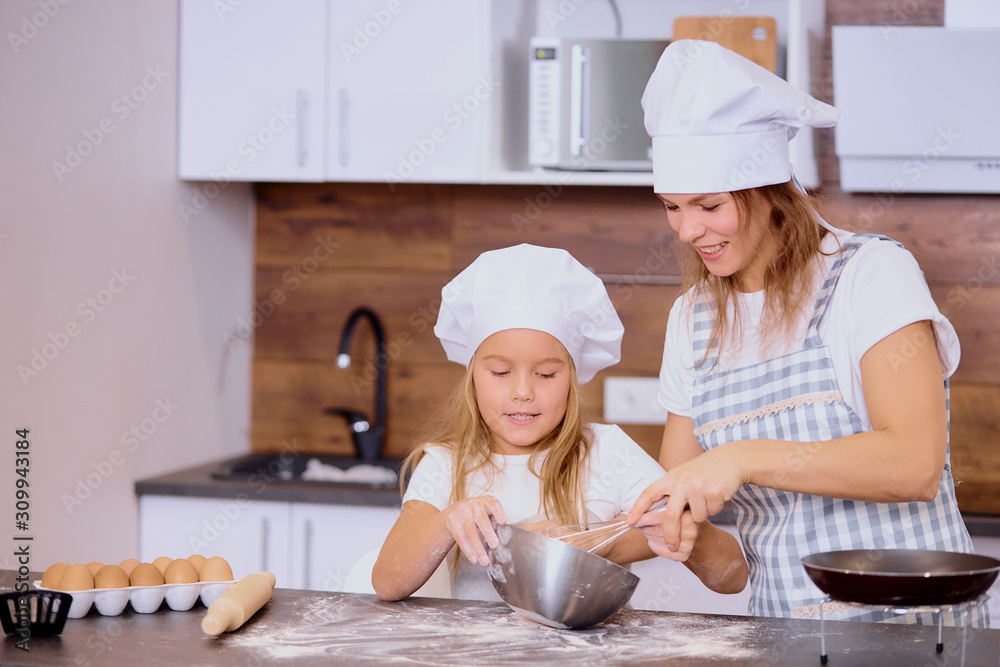 little girl mixing flour in bowl with mother in kitchen, wearing apron and caps for cooking, family teamwork little girl mixing flour in bowl with mother in kitchen, wearing apron and caps for cooking, family teamwork