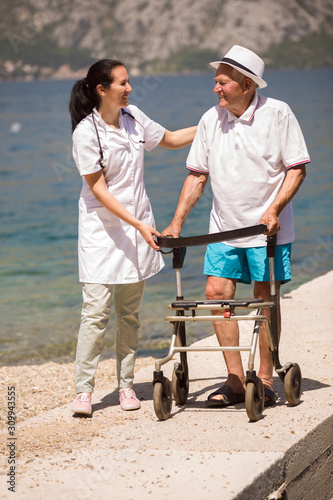 Nurse helping elderly senior man. Senior man using a walker with caregiver near the sea at summer.