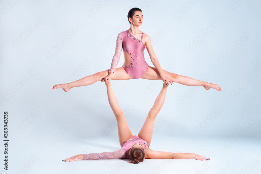 Two flexible girls gymnasts with pigtails, in pink leotards are performing splits using support while posing isolated on white background. Close-up. Two flexible girls gymnasts with pigtails, in pink leotards are performing splits using support while posing isolated on white background. Close-up.
