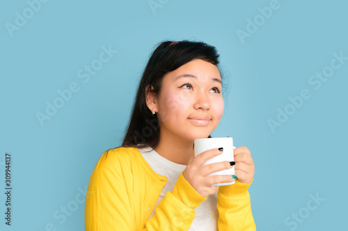 Asian teenager's portrait isolated on blue studio background. Beautiful female brunette model with long hair. Concept of human emotions, facial expression, sales, ad. Drinking coffee or tea.