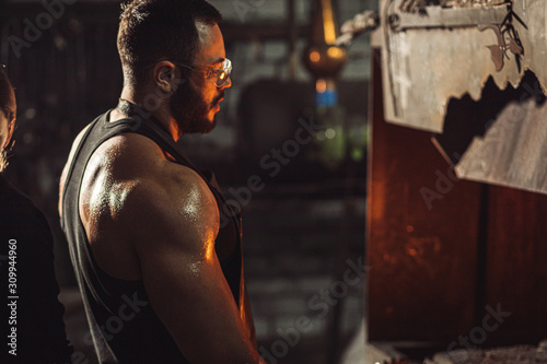 Portrait of a professional brutal caucasian blacksmith with strong muscles, side view on man opposite of fire