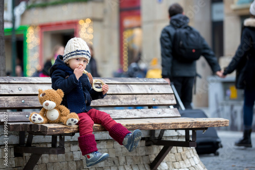 Fashion toddler boy with teddy bear toy, eating traditional Czech sweet pastry, called trdlo in the city center