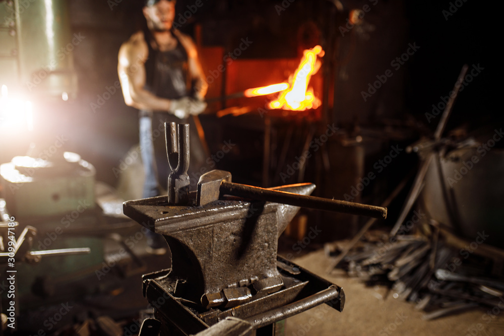 close-up of hammer and other instruments, equipment for forging, blacksmithing. muscular young man near furnace in the background