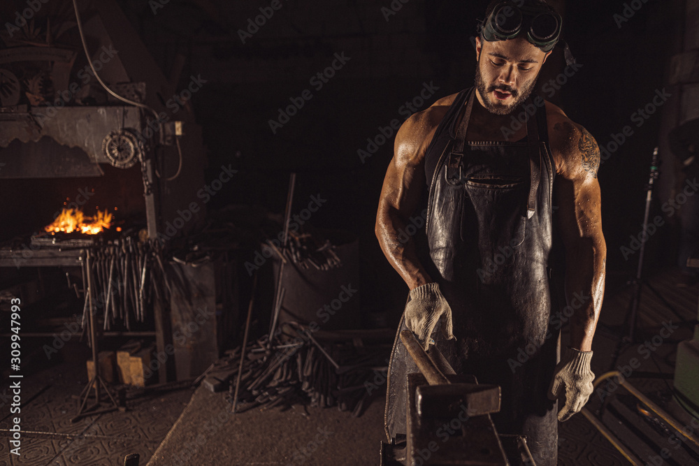 confident good-lloking artisan in leather brown apron or uniform, young caucasian forger with beard wearing gloves